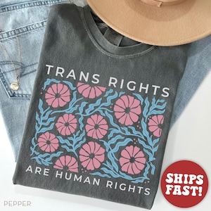 May include: A dark gray t-shirt with a floral design in pink and blue. The text "Trans Rights Are Human Rights" is printed on the shirt.