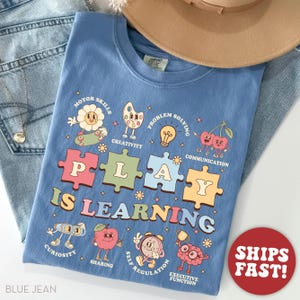 May include: A blue t-shirt with the words "PLAY IS LEARNING" in colorful puzzle piece letters. The shirt features cartoon illustrations of a flower, paint palette, lightbulb, cherries, robot, apple, clock, and brain. A tan hat and blue jeans are in the background.
