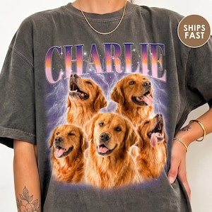 May include: A charcoal gray short-sleeve t-shirt featuring a graphic print of five golden retriever dogs. The name CHARLIE is printed in large, purple and orange gradient letters above the dogs, set against a purple lightning background.