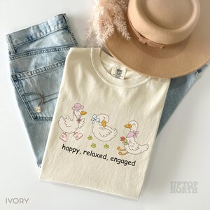 May include: Ivory-colored t-shirt with a graphic of three ducks wearing hats and bows, with the words "happy, relaxed, engaged." The shirt is paired with light blue jeans, a necklace, and a tan hat.