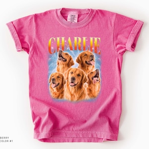 May include: A bright pink short-sleeve t-shirt featuring a graphic print of five golden retriever dogs. Above the dogs, the name CHARLIE is printed in a yellow and orange gradient font. The background of the graphic shows a blue light effect.