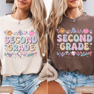 May include: Ivory and espresso-colored t-shirts with a colorful "SECOND GRADE" graphic. The design includes a smiley face, book, apple, globe, pencil, and floral accents. The shirts are paired with denim shorts.