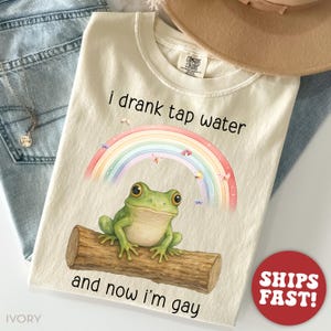 May include: A beige t-shirt with the text "I drank tap water and now I'm gay" printed on it. A rainbow with a green frog sitting on a log is above the text. The shirt is on a blue denim background.
