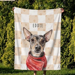 May include: A soft blanket featuring a beige and white checkered pattern with small paw prints. In the center, a detailed illustration of a dog with large ears wearing a red jacket is displayed below the name EDDIE in black text.