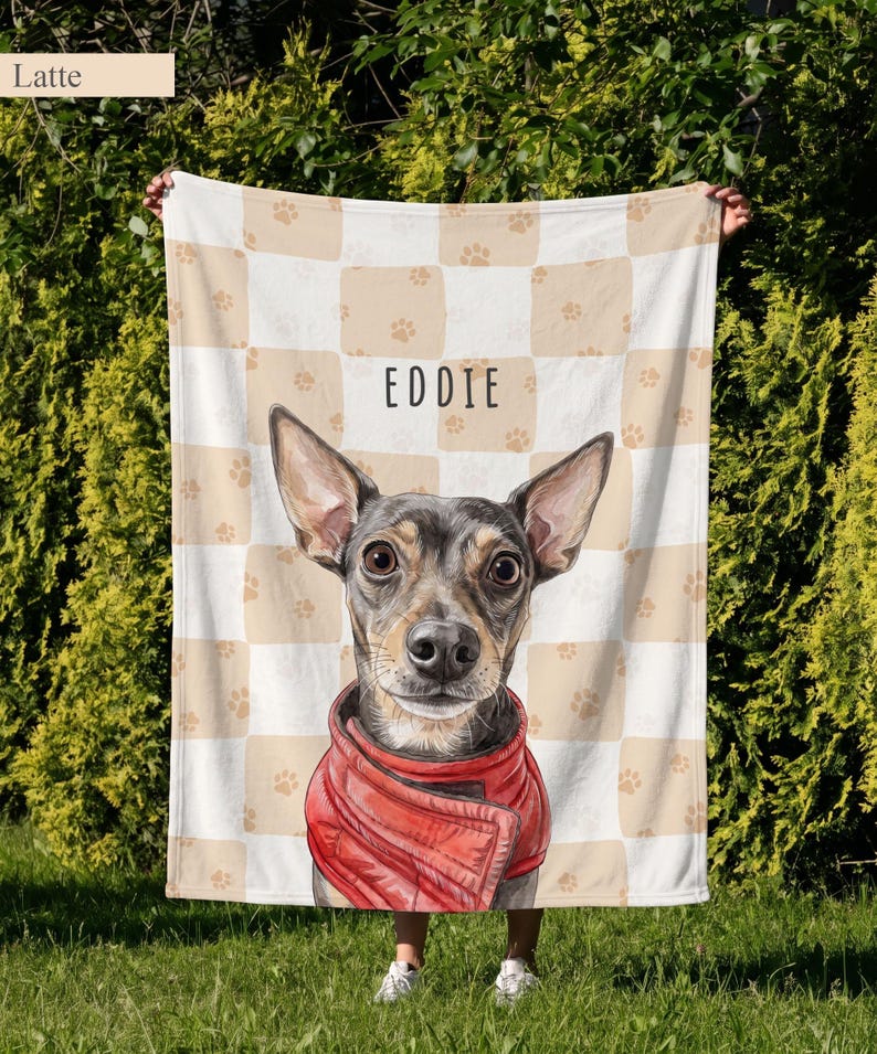 May include: A soft blanket featuring a beige and white checkered pattern with small paw prints. In the center, a detailed illustration of a dog with large ears wearing a red jacket is displayed below the name EDDIE in black text.