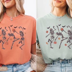 K&ouml;nnte beinhalten: Ein terrakottafarbenes T-Shirt und ein helles salbeigr&uuml;nes T-Shirt, beide mit einem Grafikdruck von drei tanzenden Skeletten, schwarzen Flederm&auml;usen und orangefarbenen Herbstbl&auml;ttern. Ein Skelett h&auml;lt einen K&uuml;rbis, ein anderes einen Eimer.