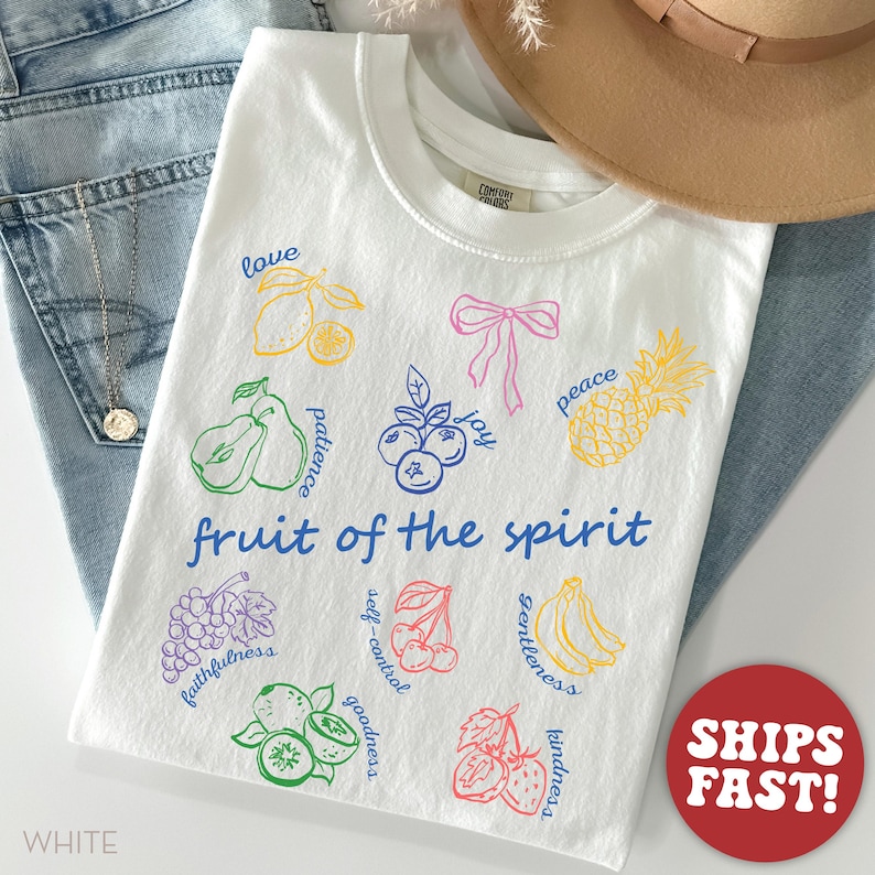 May include: White t-shirt with colorful fruit illustrations and the words "fruit of the spirit." The design includes lemons, pears, blueberries, a pineapple, grapes, cherries, bananas, and strawberries. The shirt is on a pair of blue jeans.
