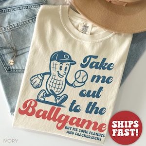 May include: A beige t-shirt with a vintage-style illustration of a peanut wearing a baseball cap and holding a baseball. The text "Take me out to the ballgame" is written in blue with a red "ballgame". The text "Buy me some peanuts and crackerjacks" is written in smaller blue text below the illustration.