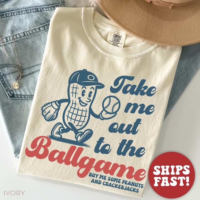 May include: A beige t-shirt with a vintage-style illustration of a peanut wearing a baseball cap and holding a baseball. The text "Take me out to the ballgame" is written in blue with a red "ballgame". The text "Buy me some peanuts and crackerjacks" is written in smaller blue text below the illustration.