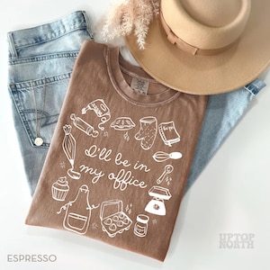 Puede incluir: Camiseta color espresso con un gr&aacute;fico blanco de utensilios de reposter&iacute;a y el texto "I'll be in my office". La camiseta se combina con vaqueros azul claro y un sombrero beige. Se ve un collar dorado.