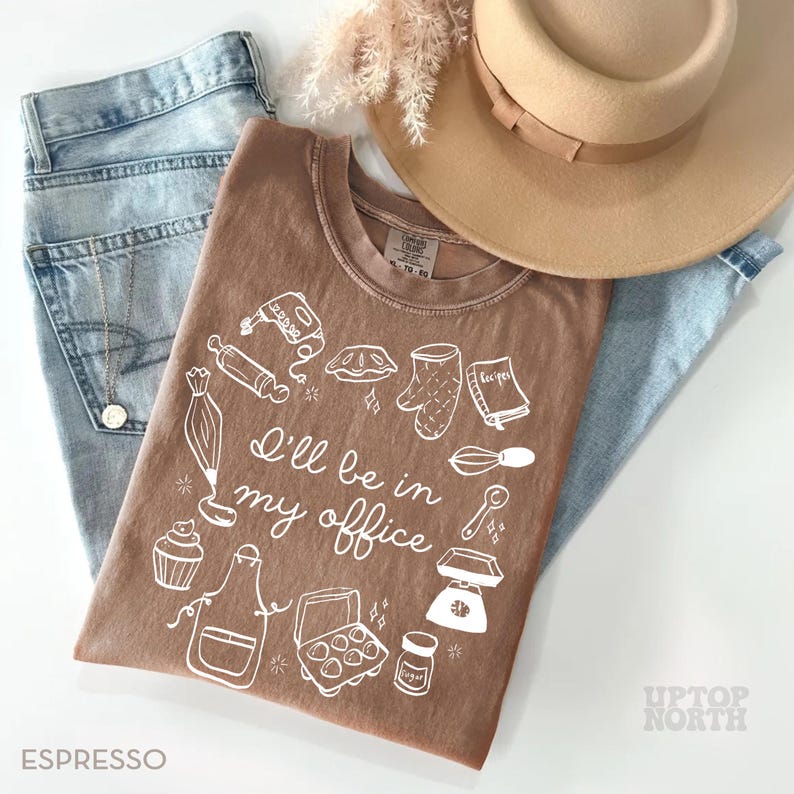 Puede incluir: Camiseta color espresso con un gr&aacute;fico blanco de utensilios de reposter&iacute;a y el texto "I'll be in my office". La camiseta se combina con vaqueros azul claro y un sombrero beige. Se ve un collar dorado.