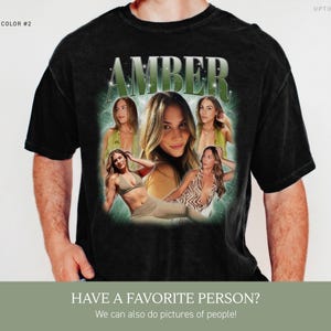 May include: A black short-sleeve t-shirt featuring a graphic print with the name AMBER in large green letters at the top. Below the name are multiple photographic images of a person in various poses, set against a glowing green background.