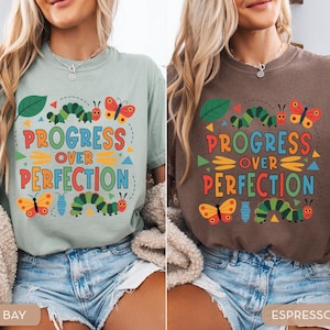 Op de afbeelding: Twee T-shirts, &eacute;&eacute;n in lichtgroen en de andere in bruin, met de tekst "PROGRESS OVER PERFECTION" in heldere kleuren. Het ontwerp bevat illustraties van rupsen, vlinders en bladeren. De shirts worden gedragen met een spijkerbroek.