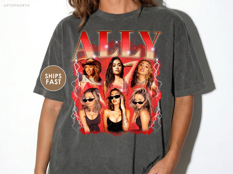 May include: Dark gray t-shirt featuring a graphic of six women with the name "ALLY" in red and gold lettering. The design includes lightning bolts and a red background. The shirt has a vintage, worn look.