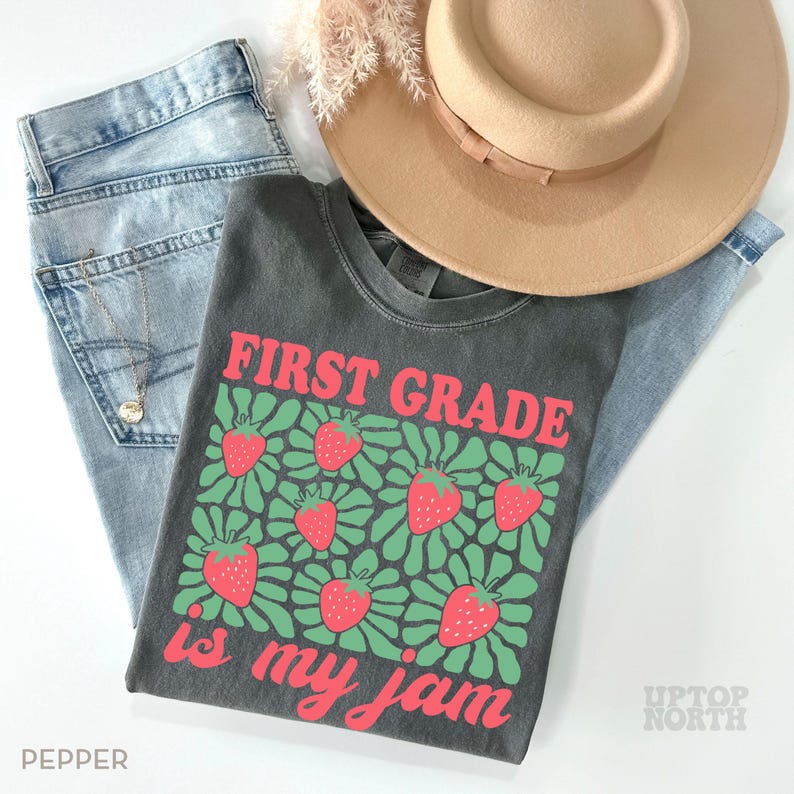 May include: A dark gray t-shirt with the text "FIRST GRADE is my jam" in red and green, featuring a strawberry design. The shirt is paired with light blue jeans and a tan hat.
