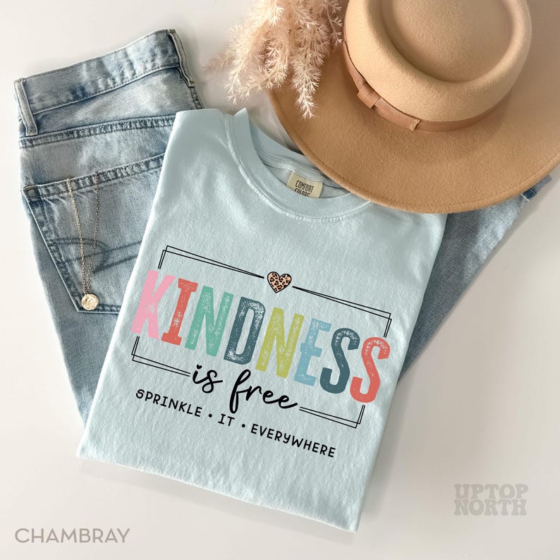 Pode incluir: Camiseta azul claro com o texto "KINDNESS is free" em cores do arco-&iacute;ris. O texto &eacute; rodeado por uma moldura preta e tem um pequeno cora&ccedil;&atilde;o com um design de estampa de leopardo acima. O texto abaixo do texto principal diz "SPRINKLE IT EVERYWHERE".