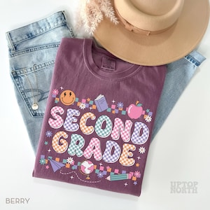 May include: A berry-colored t-shirt with the words "SECOND GRADE" in a colorful, checkered design. The shirt features school-themed graphics like an apple, book, and pencil. The shirt is paired with light blue jeans and a tan hat.