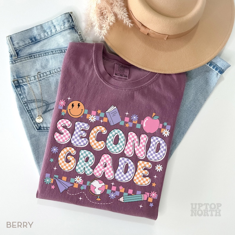 May include: A berry-colored t-shirt with the words "SECOND GRADE" in a colorful, checkered design. The shirt features school-themed graphics like an apple, book, and pencil. The shirt is paired with light blue jeans and a tan hat.