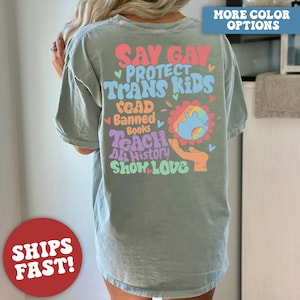 May include: A light green t-shirt with a colorful graphic design that says "Say Gay, Protect Trans Kids, Read Banned Books, Teach All History, Show Love".