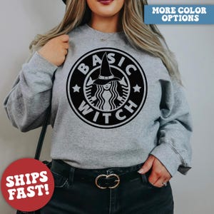 May include: A gray sweatshirt featuring a black and white graphic. The graphic is a parody of a popular coffee shop logo, with the words "BASIC WITCH" encircling a witch wearing a hat. The words "MORE COLOR OPTIONS" are in a blue box.