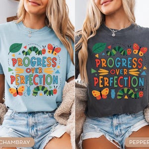 Op de afbeelding: Twee T-shirts, &eacute;&eacute;n lichtblauw en &eacute;&eacute;n donkergrijs, met de tekst "PROGRESS OVER PERFECTION" in kleurrijke letters. De shirts hebben illustraties van rupsen, vlinders en bladeren, wat een grillig ontwerp cre&euml;ert.