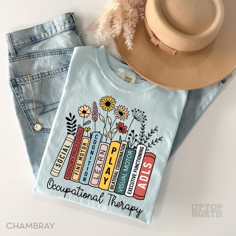 May include: Light blue t-shirt with a graphic of colorful books with words like "Social", "Sensory Processing", "Fine Motor", "Cognition", "Learn", "Play", "Bilateral Coordination", "Visual Motor", "Executive Functioning", and "ADLS" stacked on top of each other. The graphic is surrounded by flowers and the text "Occupational Therapy" is printed below the books.
