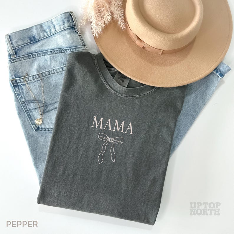 May include: A dark gray t-shirt with the word "MAMA" and a simple bow design in white. The shirt is laid out on a light blue denim background with a straw hat and a sprig of dried flowers.