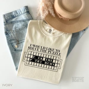May include: A cream-colored t-shirt with the text "I would like to solve the puzzle" and a black and white graphic of a crossword puzzle with the words "FCK THE" and "P TRI RCHY" inside. The shirt is on a blue denim background.