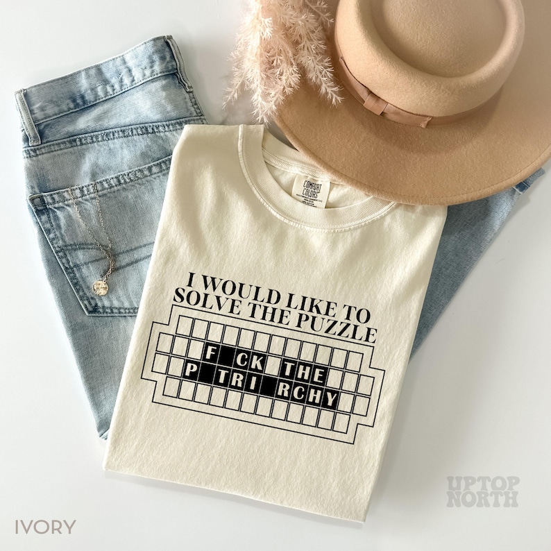May include: A cream-colored t-shirt with the text "I would like to solve the puzzle" and a black and white graphic of a crossword puzzle with the words "FCK THE" and "P TRI RCHY" inside. The shirt is on a blue denim background.