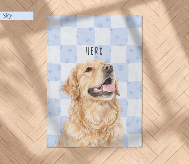 May include: A rectangular blanket featuring a light blue and white checkered pattern with small paw prints. The center displays a realistic illustration of a golden retriever with its mouth open and the text HERO printed above the dog.