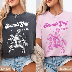 K&ouml;nnte beinhalten: Zwei Frauen tragen T-Shirts mit dem Text "Sounds Gay I'm In" und einer Grafik einer Frau, die ein Pferd reitet. Das T-Shirt links ist dunkelgrau und das T-Shirt rechts ist rosa.