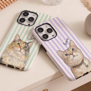 May include: Two phone cases featuring cat illustrations. One case has a green and white striped background with a cat illustration and the name "Salem". The other case has a purple and white striped background with a cat illustration and the name "Luna".