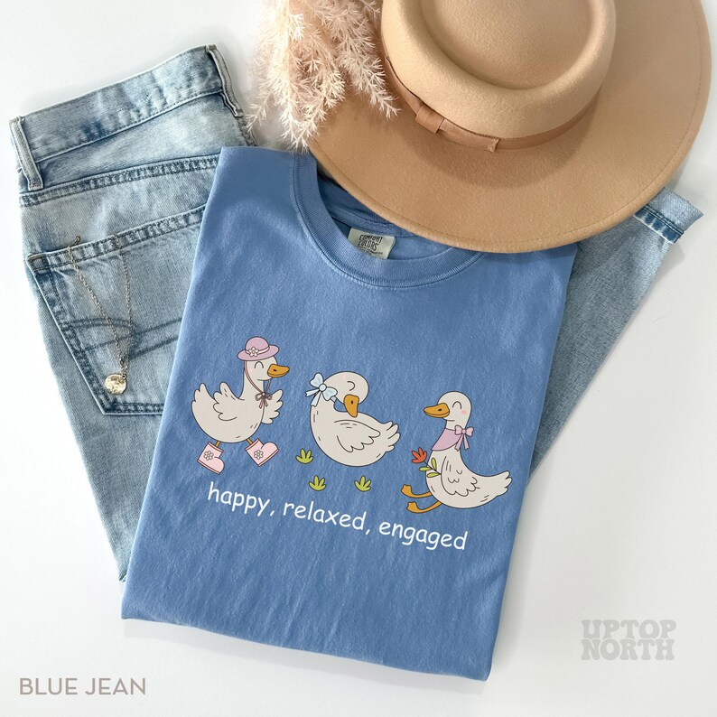 May include: A light blue t-shirt with a cartoon duck design and the words "happy, relaxed, engaged." The shirt is paired with light blue jeans and a tan hat. A necklace is visible on the jeans. The shirt is from Uptop North.