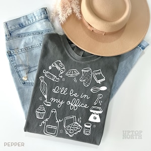Puede incluir: Una camiseta gris antracita con el texto "I'll be in my office" e ilustraciones con tem&aacute;tica de reposter&iacute;a. La camiseta se combina con unos vaqueros azul claro y un sombrero beige. Tambi&eacute;n se ve un collar y ramitas decorativas.