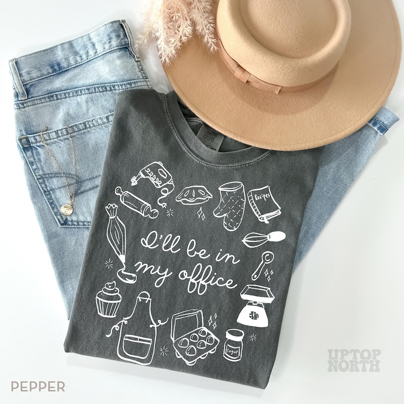 Puede incluir: Una camiseta gris antracita con el texto "I'll be in my office" e ilustraciones con tem&aacute;tica de reposter&iacute;a. La camiseta se combina con unos vaqueros azul claro y un sombrero beige. Tambi&eacute;n se ve un collar y ramitas decorativas.