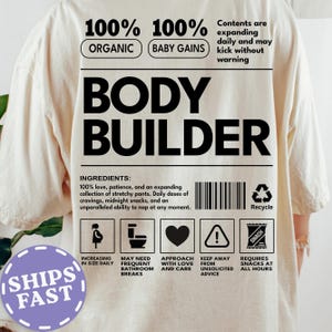 May include: A beige t-shirt with a black graphic that reads "100% Organic 100% Baby Gains Body Builder". The graphic is styled like a product label with a barcode and a recycle symbol. The label also includes a list of ingredients, which are "100% love, patience, and an expanding collection of stretchy pants. Daily doses of cravings, midnight snacks, and an unparalleled ability to nap at any moment."