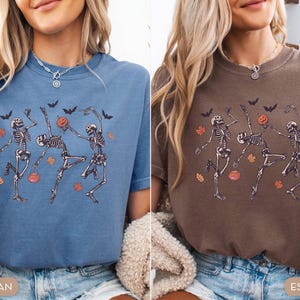K&ouml;nnte beinhalten: Ein blaues Jeans-T-Shirt und ein espressobraunes T-Shirt, beide mit einem Grafikdruck von drei tanzenden Skeletten, orangefarbenen K&uuml;rbissen, schwarzen Flederm&auml;usen und verstreuten herbstlichen Bl&auml;ttern in Orange und Gelb auf der Brust.