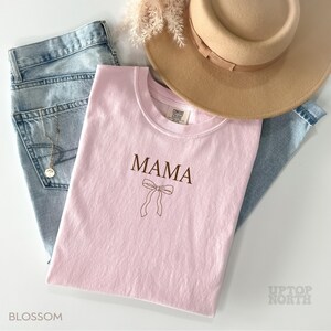 May include: Pink t-shirt with the word "MAMA" and a brown outline of a bow. The shirt is laying on a pair of blue jeans and a brown hat.