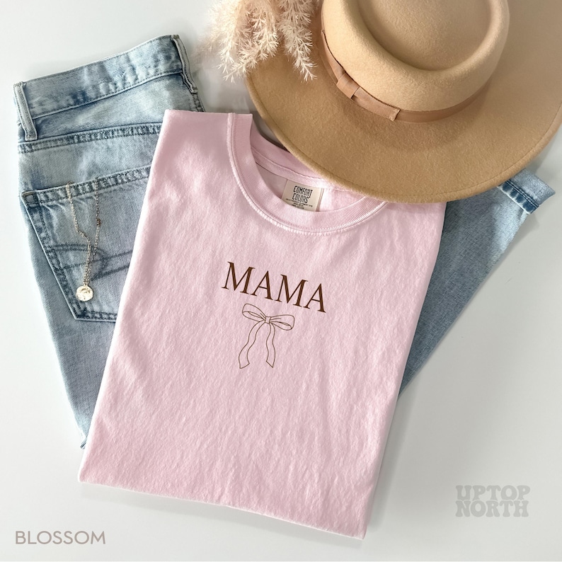 May include: Pink t-shirt with the word "MAMA" and a brown outline of a bow. The shirt is laying on a pair of blue jeans and a brown hat.