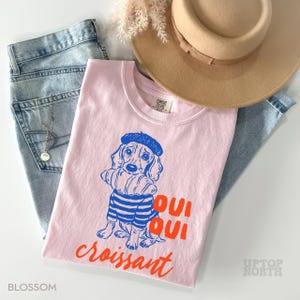 Peut inclure: Un t-shirt rose clair avec un dessin bleu et orange d'un chien portant un b&eacute;ret et tenant un croissant. Le t-shirt porte les mots "Oui Oui croissant" en orange. Le t-shirt est pli&eacute; sur un jean bleu clair et un chapeau beige.