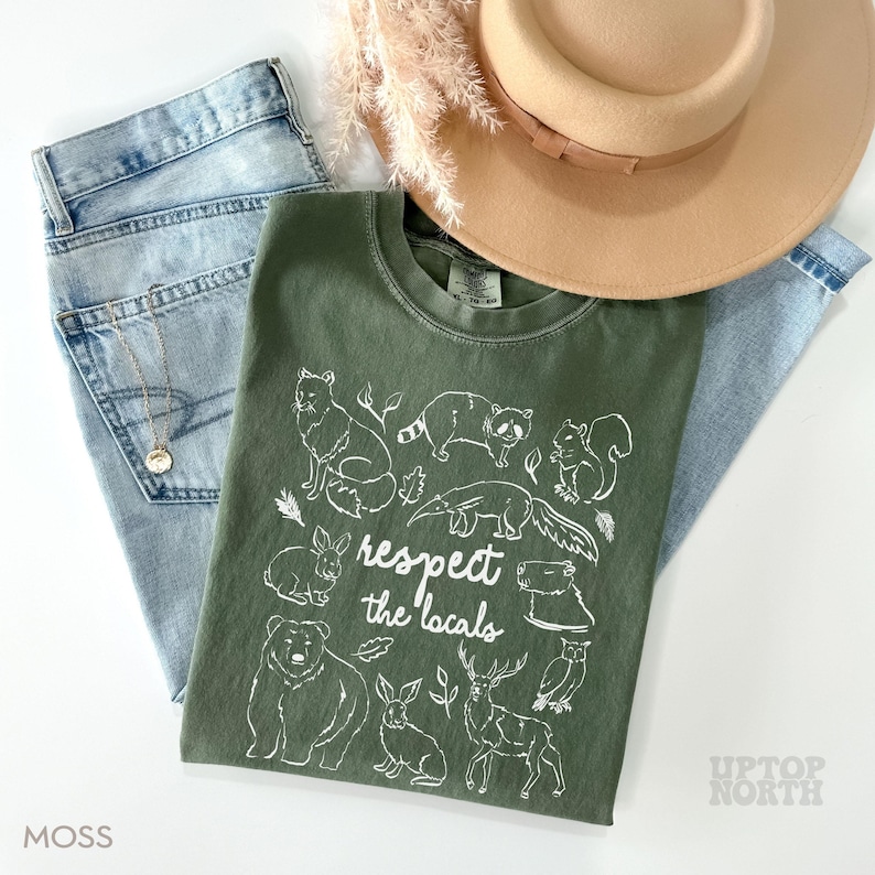 K&ouml;nnte beinhalten: Ein moosgr&uuml;nes T-Shirt mit einer wei&szlig;en Grafik verschiedener Waldtiere und dem Text "respect the locals". Das Shirt wird mit hellblauen Jeans, einem beigen Hut und einer goldenen Halskette kombiniert. Das Shirt ist ein bequemer, l&auml;ssiger Stil.