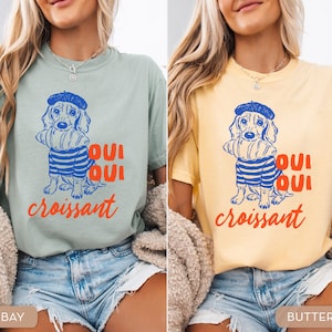 Peut inclure: Deux t-shirts, l'un vert baie et l'autre jaune beurre, chacun avec un dessin d'un teckel portant un b&eacute;ret et une chemise ray&eacute;e, tenant un croissant. Le texte "Oui Oui croissant" est imprim&eacute; en dessous du chien.