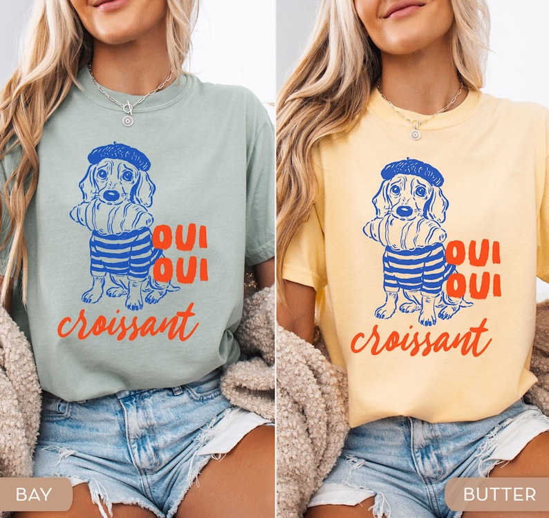 Peut inclure: Deux t-shirts, l'un vert baie et l'autre jaune beurre, chacun avec un dessin d'un teckel portant un b&eacute;ret et une chemise ray&eacute;e, tenant un croissant. Le texte "Oui Oui croissant" est imprim&eacute; en dessous du chien.