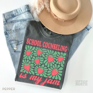 K&ouml;nnte beinhalten: Ein l&auml;ssiges Outfit mit einem grauen T-Shirt mit dem Text "SCHOOL COUNSELING is my jam" und einem Erdbeermuster. Das Shirt wird mit hellblauen Jeans und einem beigefarbenen Fedora-Hut kombiniert. Eine Halskette und eine dekorative Pflanze vervollst&auml;ndigen den Look.