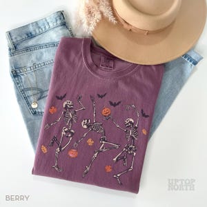K&ouml;nnte beinhalten: Ein beerenfarbenes T-Shirt mit einer Grafik von drei tanzenden Skeletten, orangefarbenen K&uuml;rbissen und schwarzen Flederm&auml;usen. Es liegt auf einer hellblauen Jeans, einem beigen Hut mit breiter Krempe und einer Goldkette. Das Etikett zeigt Comfort Colors.