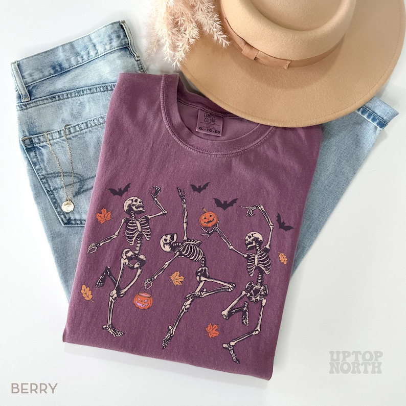 K&ouml;nnte beinhalten: Ein beerenfarbenes T-Shirt mit einer Grafik von drei tanzenden Skeletten, orangefarbenen K&uuml;rbissen und schwarzen Flederm&auml;usen. Es liegt auf einer hellblauen Jeans, einem beigen Hut mit breiter Krempe und einer Goldkette. Das Etikett zeigt Comfort Colors.