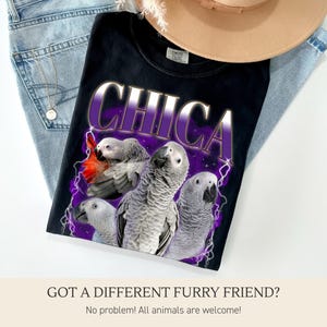 Pu&ograve; includere: T-shirt nero con la scritta "CHICA" in un design viola e bianco, con una grafica di quattro pappagalli grigi con accenti rossi. La maglietta &egrave; appoggiata su jeans blu e un cappello beige.
