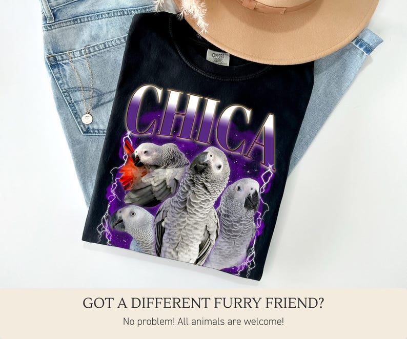 Pu&ograve; includere: T-shirt nero con la scritta "CHICA" in un design viola e bianco, con una grafica di quattro pappagalli grigi con accenti rossi. La maglietta &egrave; appoggiata su jeans blu e un cappello beige.