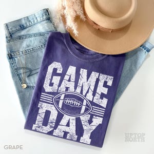 May include: A grape-coloured t-shirt with the words "GAME DAY" and a football graphic, paired with light-wash jeans and a tan hat. The shirt has a distressed look. A gold necklace is visible.