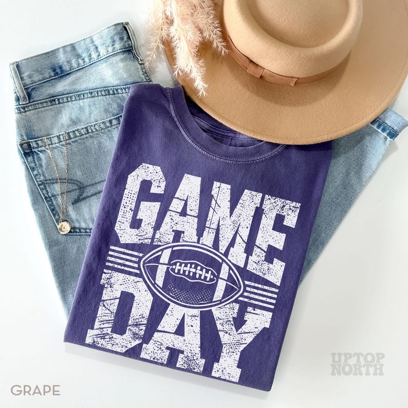 May include: A grape-coloured t-shirt with the words "GAME DAY" and a football graphic, paired with light-wash jeans and a tan hat. The shirt has a distressed look. A gold necklace is visible.